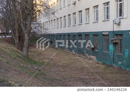 Old institutional building exterior with tiled facade and metal window grates near a barren slope, conveying an urban atmosphere of neglect and history in a quiet city environment Old institutional building exterior with tiled facade and metal window grates near a barren slope, conveying an urban atmosphere of neglect and history in a quiet city environment 140146226