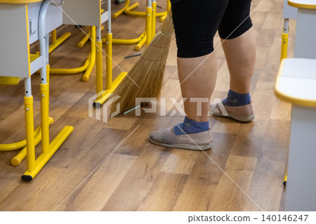A professional custodian sweeps the wooden floor of a school classroom using a traditional straw broom between rows of desks to maintain a clean learning environment for students A professional custodian sweeps the wooden floor of a school classroom using a traditional straw broom between rows of desks to maintain a clean learning environment for students 140146247
