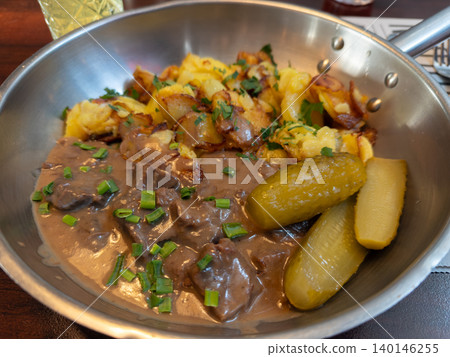 Hearty beef stew with golden roasted potatoes and pickles served in a traditional metal skillet, representing rustic European cuisine and a comforting authentic dining experience Hearty beef stew with golden roasted potatoes and pickles served in a traditional metal skillet, representing rustic European cuisine and a comforting authentic dining experience 140146255