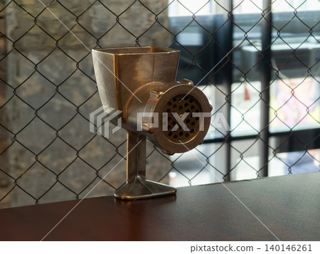 A vintage metal meat grinder rests on a rustic wooden table against a wire mesh background, creating a classic atmosphere ideal for culinary themes or industrial cafe decor 140146261