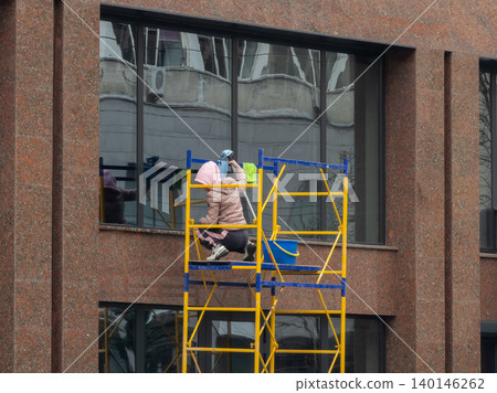 A professional worker cleans a glass office facade while standing on yellow metal scaffolding at an exterior building site to showcase expert janitorial maintenance services today 140146262