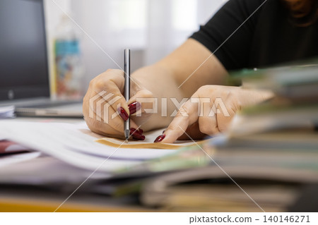 A professional person with vibrant red nail polish writes carefully with a pen on important office documents to convey organized administrative tasks and meticulous corporate planning 140146271