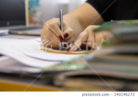 Person writing with a pen on paper at a cluttered desk, conveying focus and administrative work tasks in a productive professional business office environment with a close up view 140146272