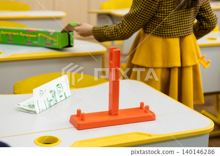 An orange plastic educational tool sits on a classroom desk near a student holding a game box, depicting creative STEM learning and engineering activities in a modern primary school An orange plastic educational tool sits on a classroom desk near a student holding a game box, depicting creative STEM learning and engineering activities in a modern primary school 140146286