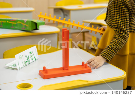 A young child plays with an orange plastic math scale toy on a classroom desk to learn arithmetic principles, illustrating hands-on educational tools for early school development 140146287