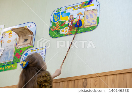 Schoolgirl pointing with a wooden pointer at a Ukrainian educational wall poster, demonstrating a student engaging in learning about national symbols within a classroom environment Schoolgirl pointing with a wooden pointer at a Ukrainian educational wall poster, demonstrating a student engaging in learning about national symbols within a classroom environment 140146288