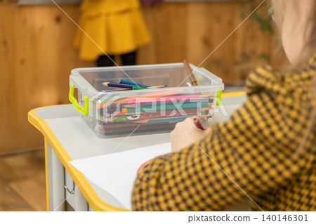 A young child is drawing with colored pencils at a wooden desk in a bright classroom, showcasing a creative learning environment where students develop artistic skills today A young child is drawing with colored pencils at a wooden desk in a bright classroom, showcasing a creative learning environment where students develop artistic skills today 140146301