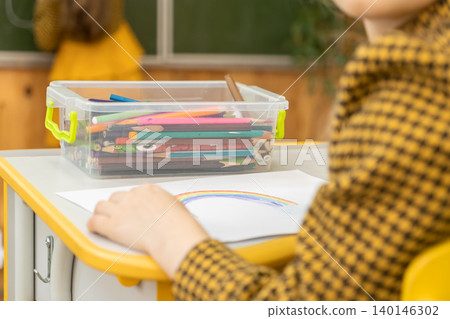 A young student draws a colorful rainbow on white paper using pencils from a clear plastic organizer on a yellow school desk inside a bright and modern classroom learning environment A young student draws a colorful rainbow on white paper using pencils from a clear plastic organizer on a yellow school desk inside a bright and modern classroom learning environment 140146302