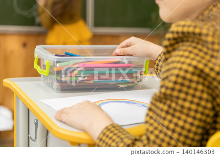 A young student selects a vibrant colored pencil from a plastic container at their desk to draw a rainbow on paper, capturing creative atmosphere and expression in the classroom A young student selects a vibrant colored pencil from a plastic container at their desk to draw a rainbow on paper, capturing creative atmosphere and expression in the classroom 140146303