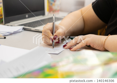 Person writing with a black pen on a small white paper note at a bright desk, conveying professional focus and active office planning within a modern corporate remote work environment 140146306