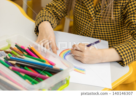 A young child is drawing a vibrant rainbow with colored pencils at a bright classroom desk to foster creative expression and artistic learning during early childhood development A young child is drawing a vibrant rainbow with colored pencils at a bright classroom desk to foster creative expression and artistic learning during early childhood development 140146307