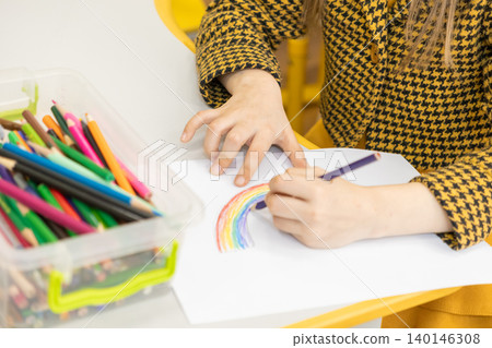 A young child draws a vibrant colorful rainbow with pencils on white paper at a desk, fostering creativity, imagination, and learning during an artistic activity in a classroom A young child draws a vibrant colorful rainbow with pencils on white paper at a desk, fostering creativity, imagination, and learning during an artistic activity in a classroom 140146308