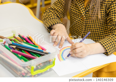 A young child is carefully drawing a colorful rainbow with a purple pencil at a school desk while creating a creative artistic project during a fun lesson in a bright classroom A young child is carefully drawing a colorful rainbow with a purple pencil at a school desk while creating a creative artistic project during a fun lesson in a bright classroom 140146309