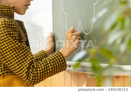 A young school girl draws on a green chalkboard using white chalk to capture a creative moment of learning and artistic expression in a brightly lit classroom for education A young school girl draws on a green chalkboard using white chalk to capture a creative moment of learning and artistic expression in a brightly lit classroom for education 140146313