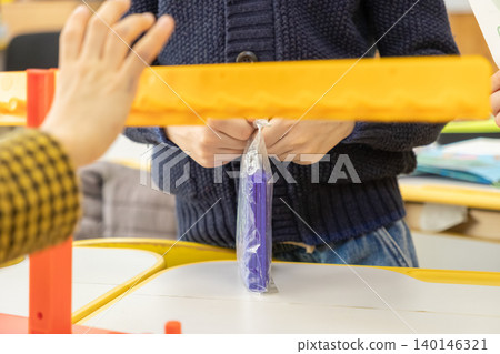 A student carefully unpacks a colorful plastic toy set at a bright school desk while a curious peer watches the collaborative elementary educational activity with great interest 140146321