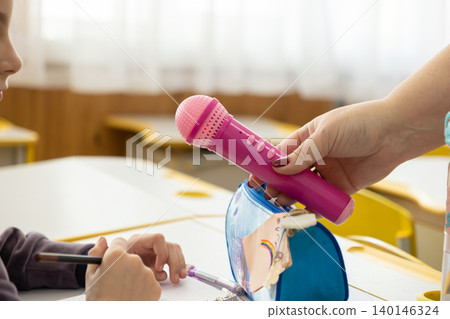 Teacher offering a pink toy microphone to a student at a classroom desk, fostering an interactive and engaging atmosphere during a creative school activity for young pupils to learn Teacher offering a pink toy microphone to a student at a classroom desk, fostering an interactive and engaging atmosphere during a creative school activity for young pupils to learn 140146324