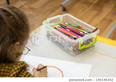 A young girl is drawing with vibrant colored pencils on white paper at her school desk while engaging in a creative and artistic educational activity inside a bright classroom A young girl is drawing with vibrant colored pencils on white paper at her school desk while engaging in a creative and artistic educational activity inside a bright classroom 140146342