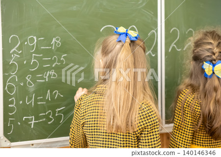 Two primary school girls writing multiplication math problems on a green blackboard in a classroom, representing elementary education, learning, academic growth, and intelligence 140146346