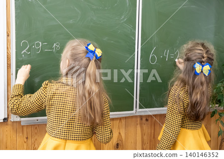 Two young schoolgirls in yellow dresses solve complex math equations with white chalk on a large green classroom blackboard, representing academic learning and school activities 140146352