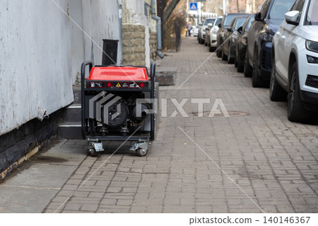 A red portable electric power generator sits on a paved city sidewalk near parked cars, depicting an emergency energy solution during a sudden blackout for homes or businesses A red portable electric power generator sits on a paved city sidewalk near parked cars, depicting an emergency energy solution during a sudden blackout for homes or businesses 140146367