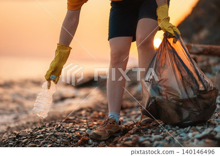 Earth day. Cleanup garbage on the ocean coast. A volunteer with polyethielene bag picking up a plastic bottle on the beach. Close-up. Sunset in the background. The concept of conservation of ecology Earth day. Cleanup garbage on the ocean coast. A volunteer with polyethielene bag picking up a plastic bottle on the beach. Close-up. Sunset in the background. The concept of conservation of ecology 140146496