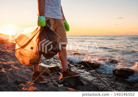A male volunteer holding two polyethilene bag with garbage. Close up. In the background the sea and the sunset. Copy space. The concept of environmental protection and Earth day 140146502