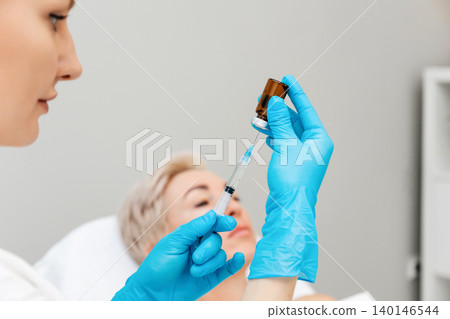 Cosmetologist is taking medicine into a syringe for a rejuvenating injection. In the background lies a woman on the procedure. Concept of professional cosmetology Cosmetologist is taking medicine into a syringe for a rejuvenating injection. In the background lies a woman on the procedure. Concept of professional cosmetology 140146544