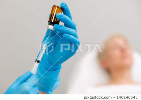 Cosmetologist's hands is taking medicine into a syringe for a rejuvenating injection. In the background lies a woman on the procedure. Concept of professional cosmetology Cosmetologist's hands is taking medicine into a syringe for a rejuvenating injection. In the background lies a woman on the procedure. Concept of professional cosmetology 140146545