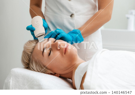 Professional care. A cosmetologist in blue gloves does an ultrasound cleaning of a woman's face. Cleansing and rejuvenating procedure in a beauty salon. Side view Professional care. A cosmetologist in blue gloves does an ultrasound cleaning of a woman's face. Cleansing and rejuvenating procedure in a beauty salon. Side view 140146547