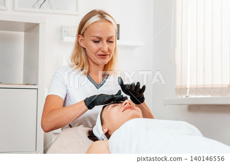 Cosmetology and lifting. Professional beautician in latex gloves gives a facial massage to the client in salon. Concept of beauty procedures Cosmetology and lifting. Professional beautician in latex gloves gives a facial massage to the client in salon. Concept of beauty procedures 140146556