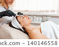 Professional cosmetology. Beautician in rubber gloves cleans the client's forehead using a hydra-peeling device. Side view. Concept of beauty procedures 140146558