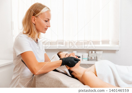 Professional beautician in protective gloves gives a rejuvenation facial massage to the client in salon. Concept of cosmetology and beauty procedure 140146559