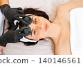 Professional beautician gives a facial rejuvenation massage with jade stones to the client in salon. Close up of female face. Top view. Concept of cosmetology and beauty procedures 140146561