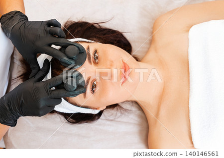 Professional beautician gives a facial rejuvenation massage with jade stones to the client in salon. Close up of female face. Top view. Concept of cosmetology and beauty procedures 140146561