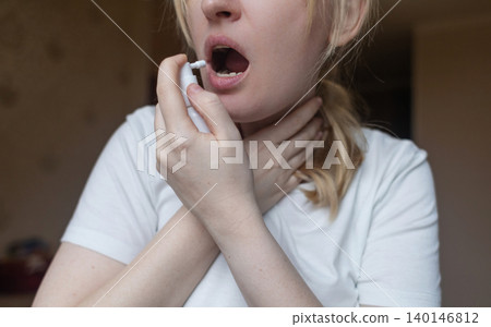 Blonde woman using throat spray to relieve pain. Close up of female treating sore throat or seasonal illness with medication at home. Blonde woman using throat spray to relieve pain. Close up of female treating sore throat or seasonal illness with medication at home. 140146812