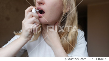 Blonde woman using throat spray to relieve pain. Close up of female treating sore throat or seasonal illness with medication at home. Blonde woman using throat spray to relieve pain. Close up of female treating sore throat or seasonal illness with medication at home. 140146814