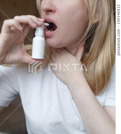 Blonde woman using throat spray to relieve pain. Close up of female treating sore throat or seasonal illness with medication at home. Blonde woman using throat spray to relieve pain. Close up of female treating sore throat or seasonal illness with medication at home. 140146815