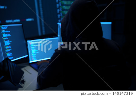 Hacker in Hoodie Working at Multiple Computer Monitors 140147319