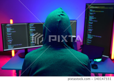 Hooded hacker coding at dual monitors in neon lit room Hooded hacker coding at dual monitors in neon lit room 140147331