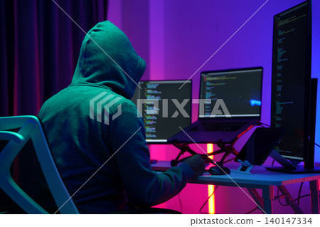 Hooded Hacker Working at Multiple Monitors in Dark Room 140147334