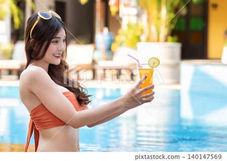 Sexy young asian woman in swimsuits in summer. beautiful woman in orange bikini with sunglasses and tropical drink sitting in swimming pool. 140147569
