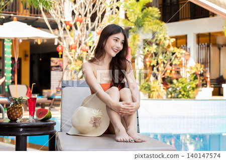 Sexy young asian woman in swimsuits in summer. beautiful woman in orange bikini with tropical drink sitting on sofa by swimming pool. Sexy young asian woman in swimsuits in summer. beautiful woman in orange bikini with tropical drink sitting on sofa by swimming pool. 140147574
