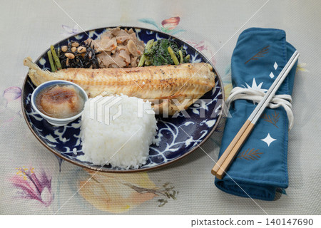 A grilled Shima Hokke fillet sits on a deep blue patterned plate, paired with a mound of white rice and a small dipping bowl. Three traditional sides hijiki seaweed, simmered roots, and greens line A grilled Shima Hokke fillet sits on a deep blue patterned plate, paired with a mound of white rice and a small dipping bowl. Three traditional sides hijiki seaweed, simmered roots, and greens line 140147690