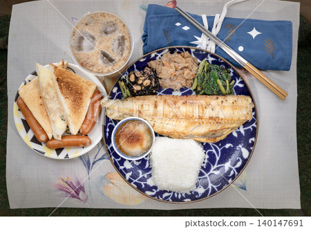 A blue patterned plate holds a grilled fish fillet, white rice, grated radish, and three sides: hijiki seaweed, simmered roots, and greens. To the left, a smaller plate has toast triangles and three A blue patterned plate holds a grilled fish fillet, white rice, grated radish, and three sides: hijiki seaweed, simmered roots, and greens. To the left, a smaller plate has toast triangles and three 140147691