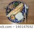 A grilled golden-brown fish fillet rests diagonally on a patterned deep-blue ceramic plate over a wooden table. It is accompanied by a square-molded portion of white rice, a small condiment bowl and 140147692