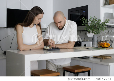 Attractive couple in cozy homewear playing chess in modern interior at home Attractive couple in cozy homewear playing chess in modern interior at home 140147784