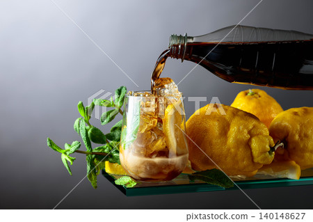 Cola is poured from a bottle into a glass with lemon wedges, ice and mint. Cola is poured from a bottle into a glass with lemon wedges, ice and mint. 140148627