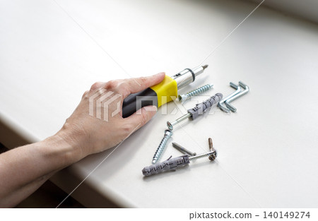 Hand holding interchangeable screwdriver near screws with plastic dowels and hex keys on white windowsill, PVC window repair or adjustment process 140149274