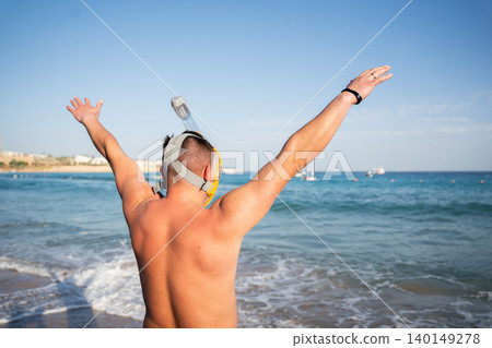 Caucasian man wearing a full face snorkeling mask stands on a beach in Sharm El Sheikh Egypt with arms raised toward the Red Sea. Beach sea vacation travel resort lifestyle Caucasian man wearing a full face snorkeling mask stands on a beach in Sharm El Sheikh Egypt with arms raised toward the Red Sea. Beach sea vacation travel resort lifestyle 140149278