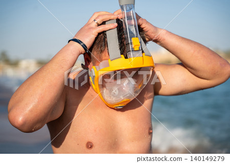 Caucasian man adjusts a full face snorkeling mask on a beach in Sharm El Sheikh Egypt with Red Sea water in the background. Beach sea vacation resort lifestyle Caucasian man adjusts a full face snorkeling mask on a beach in Sharm El Sheikh Egypt with Red Sea water in the background. Beach sea vacation resort lifestyle 140149279
